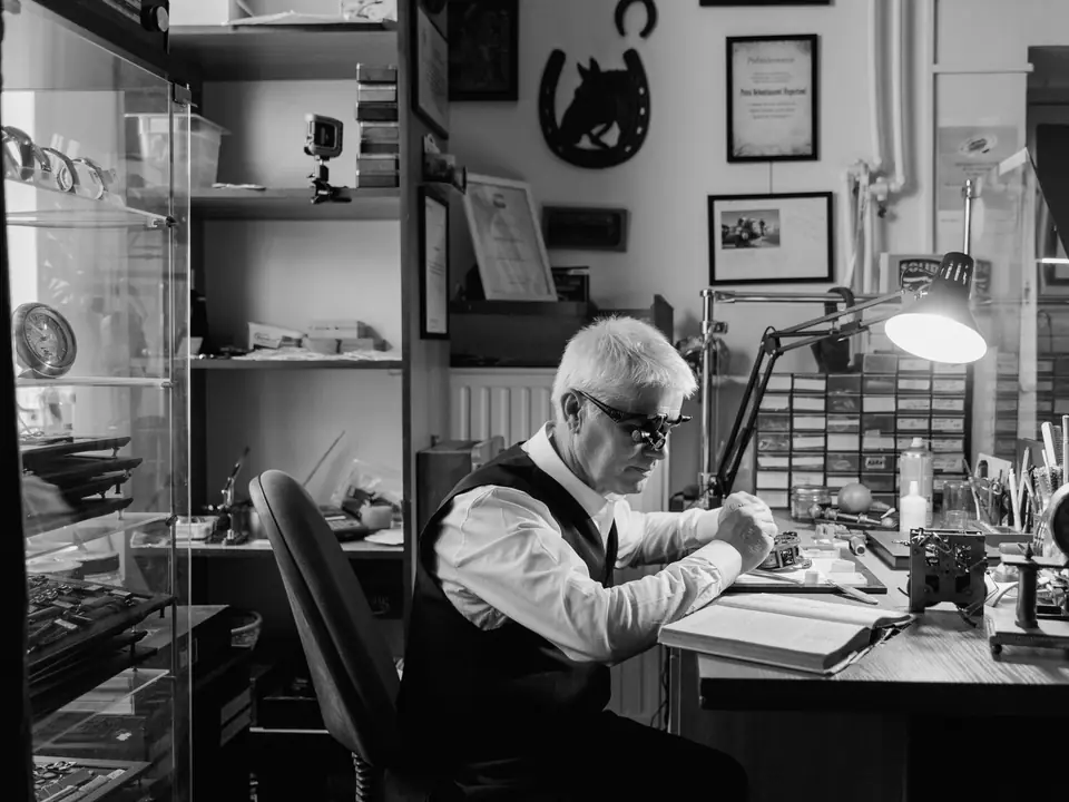A gray-haired man in a vest works at a desk under a lamp in a small shop lined with shelves.