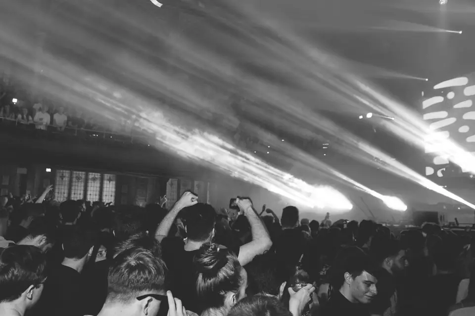 A crowd of young people dances with raised arms under sweeping stage light beams.
