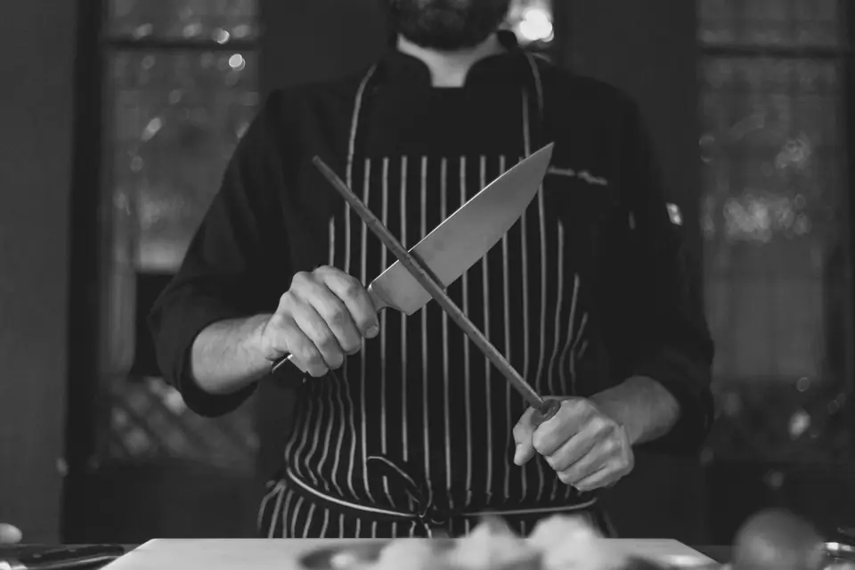 A cook in a striped apron hones a chef’s knife against a sharpening steel held upright.