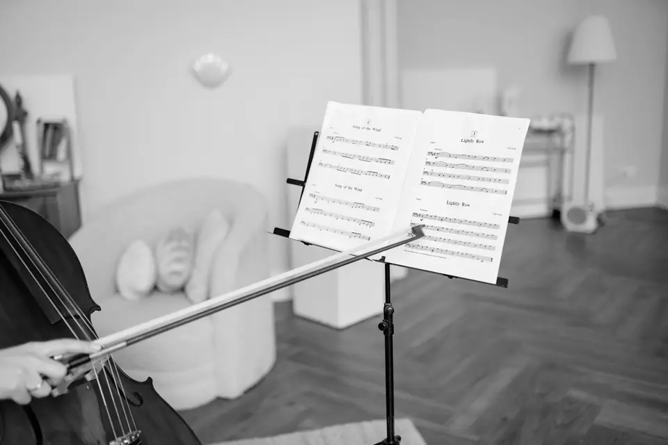 A music stand holds open sheet music in a bright living room while a cellist plays at the left edge.
