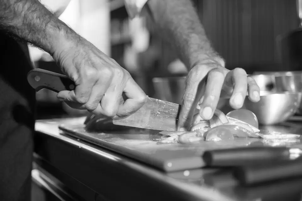 A cook’s hands slice food with a chef’s knife on a cutting board in a professional kitchen.
