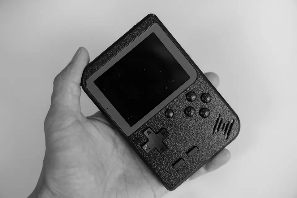 An open hand holds a small dark handheld game device with a screen, direction pad and four buttons.