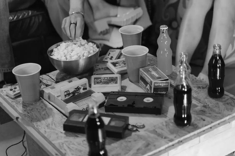 A table holds game cartridges, a console, videotapes, popcorn, cups and soda bottles during a gathering.