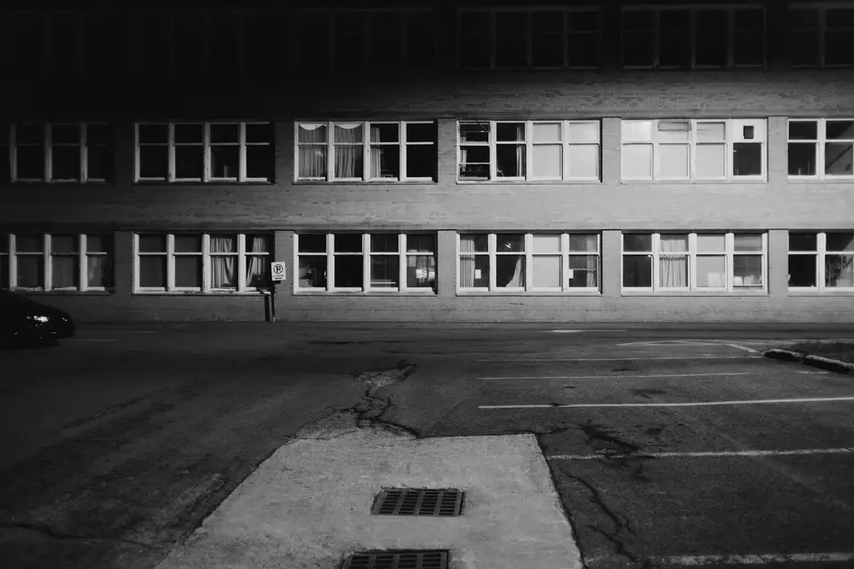 A single person walks past a long, two-story building with rows of windows beside an empty parking lot.