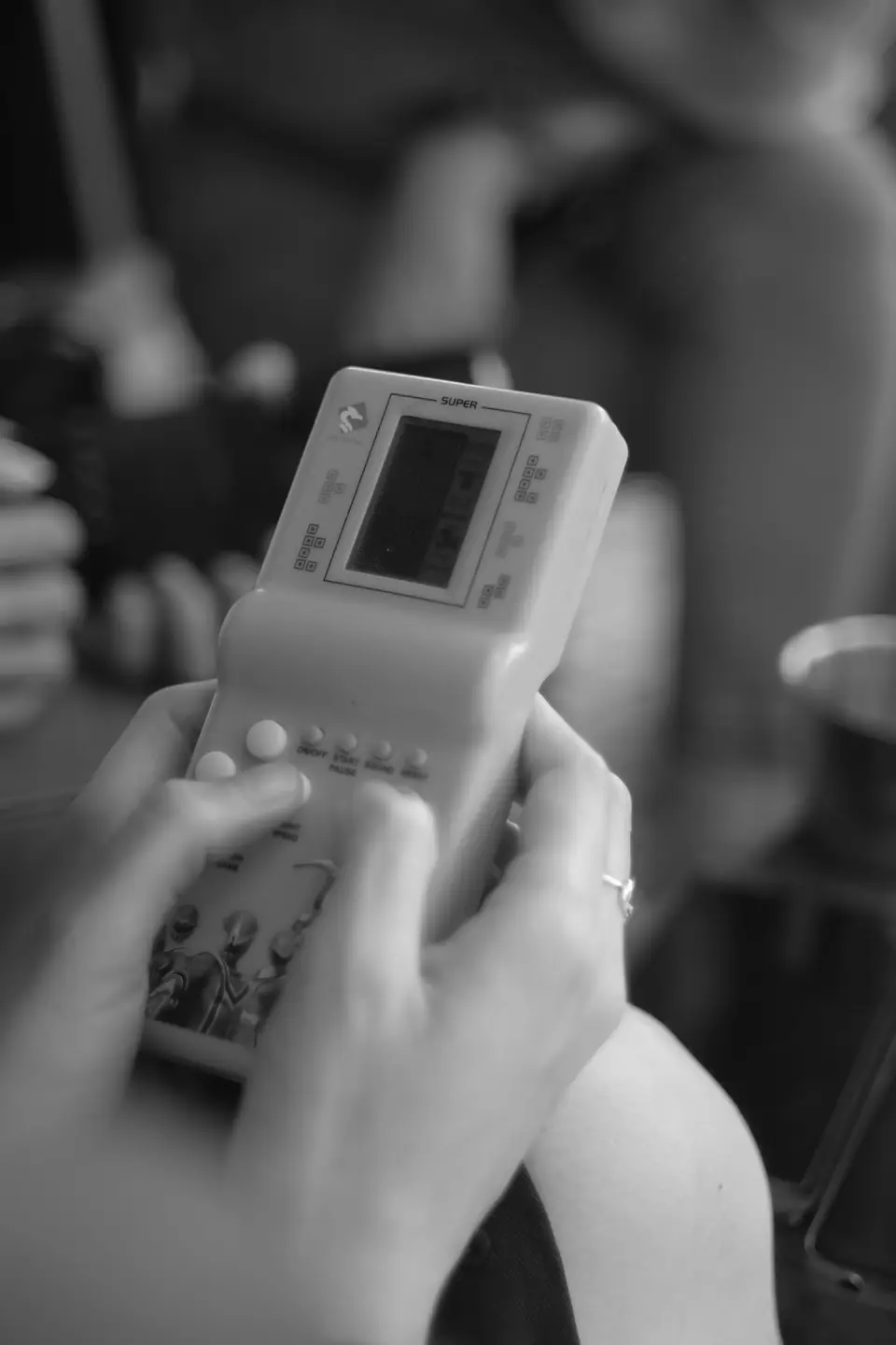 A person holds a small handheld brick game with a monochrome screen, the background softly blurred.