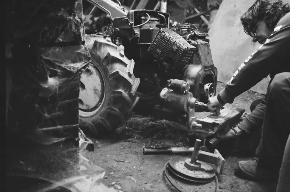 A man crouches and works on an opened tractor engine next to a large rear tire.