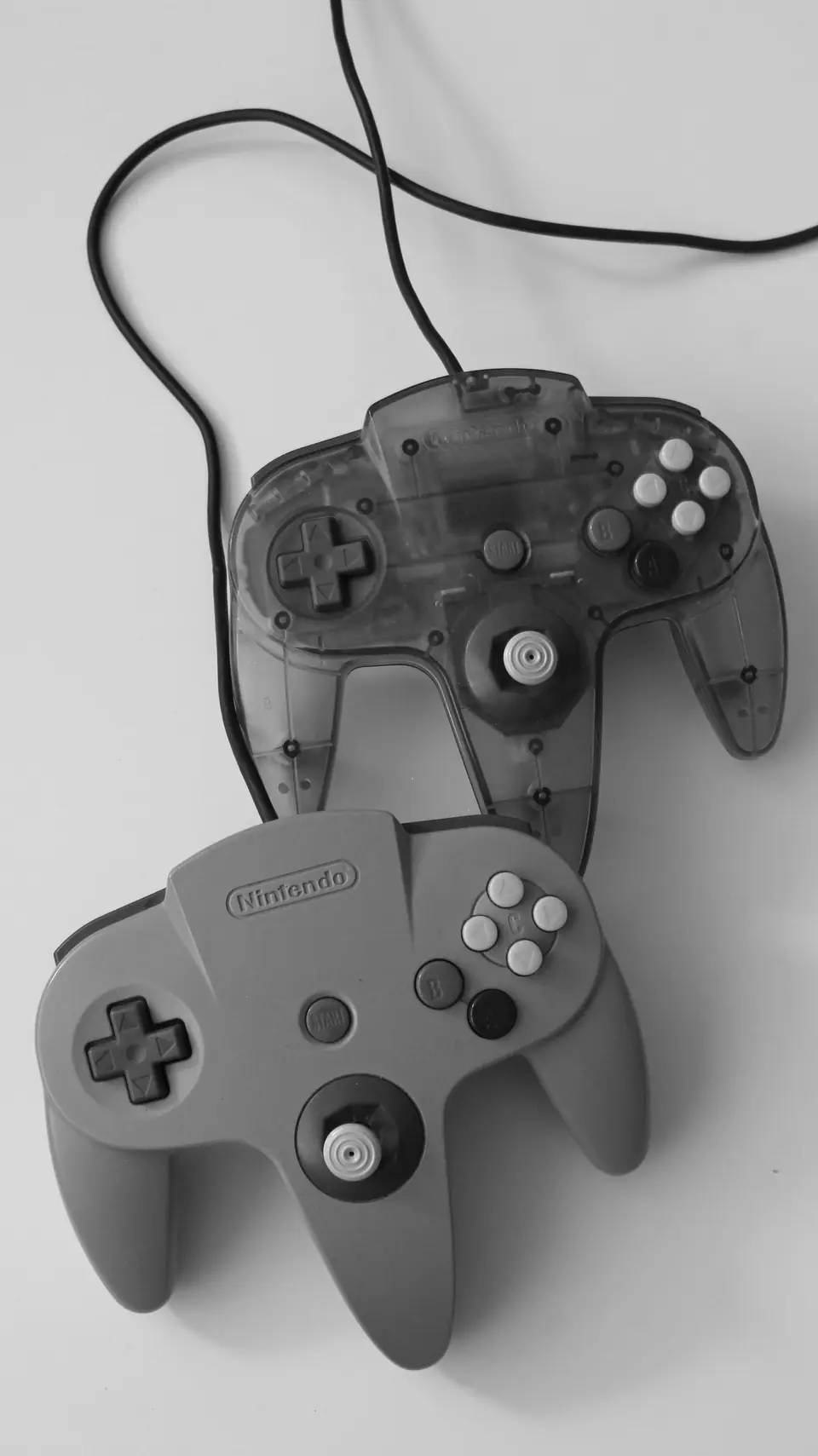 Two three-pronged game controllers, one gray and one translucent, lie on a white surface with cords.