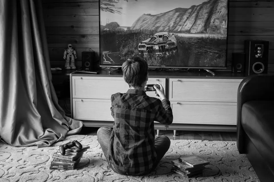 A child in a plaid shirt sits on a rug holding a game controller in front of a large television.