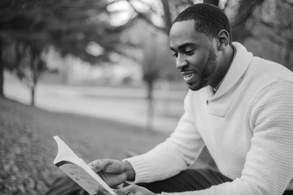 A smiling man in a white shawl-collar sweater reads a paperback book outdoors on the grass.