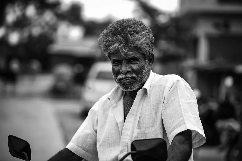 A gray-haired man with a beard, in a white shirt, sits on a motorbike on a street.