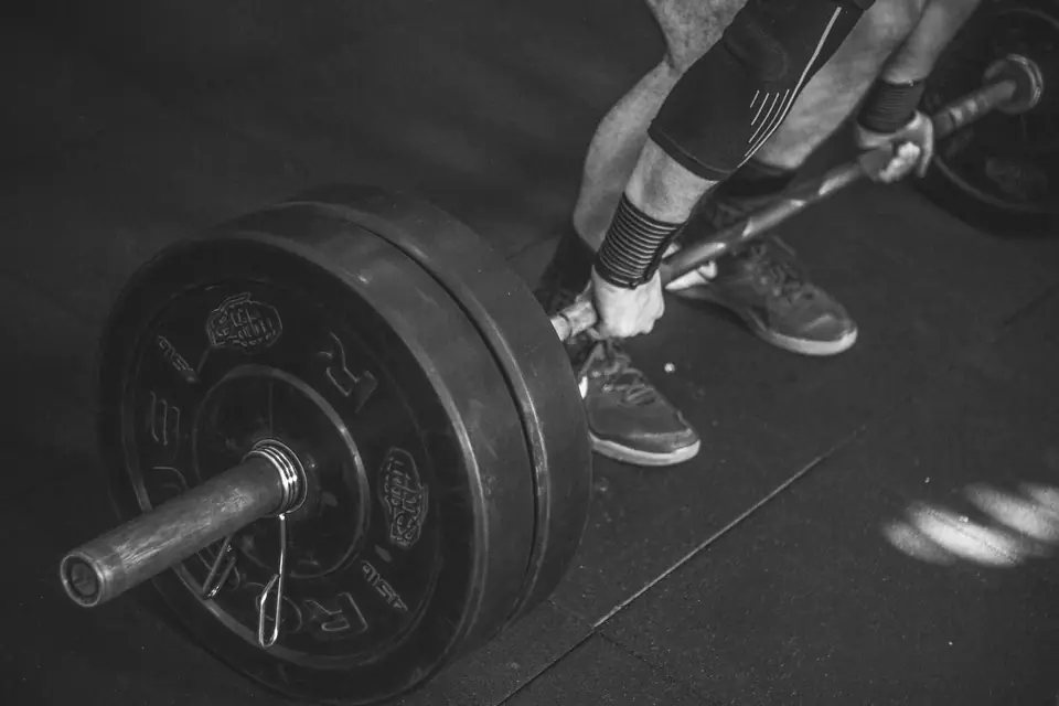 A lifter in knee sleeves grips a barbell loaded with heavy plates on a dark gym floor.