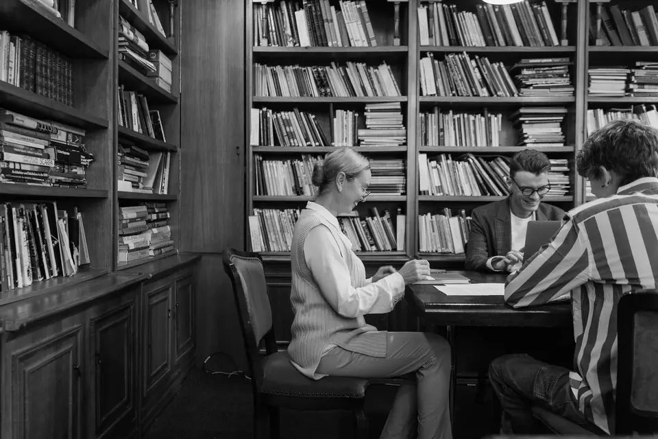 Three people sit at a table talking in a room lined with tall wooden bookshelves.