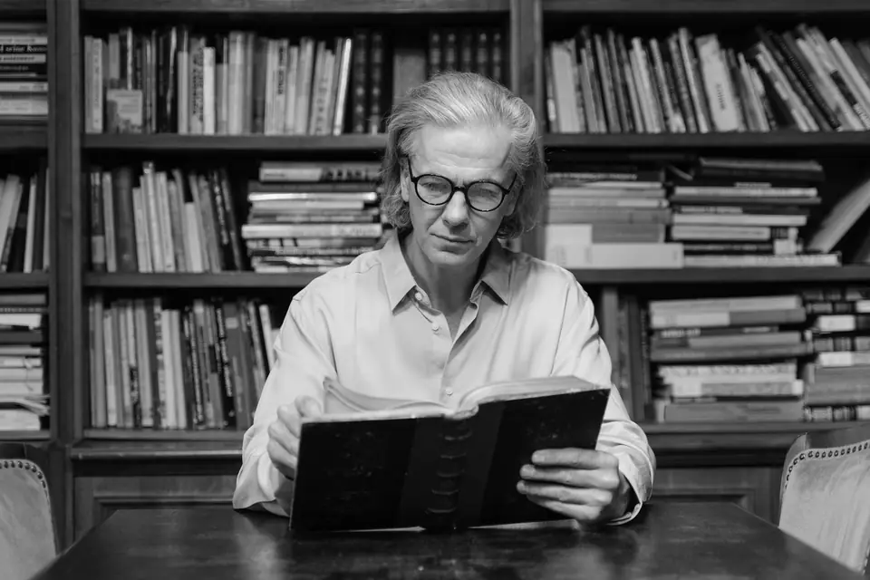 A man with long gray hair and glasses reads a large book at a table before crowded bookshelves.