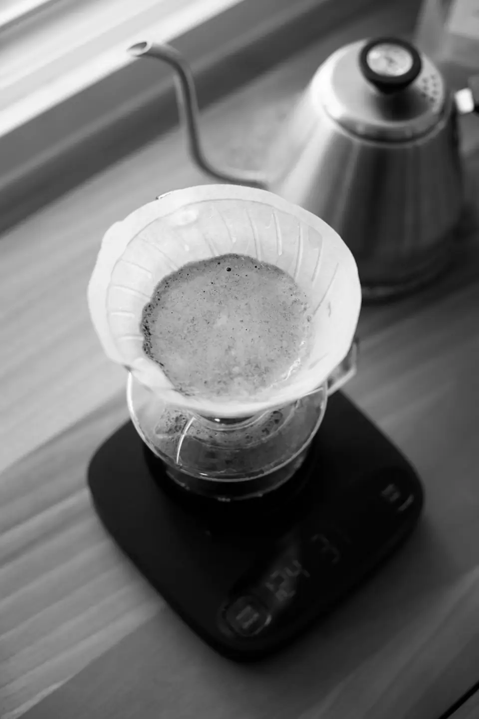 A pour-over coffee cone with a paper filter sits on a digital scale beside a gooseneck kettle.