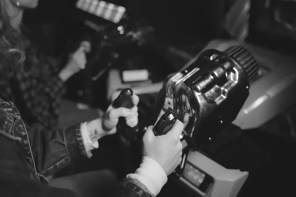 Two people hold joystick controls on side-by-side arcade cabinets in a dim room.