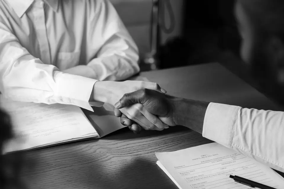 Two people in white shirts shake hands across a table holding printed documents and a pen.